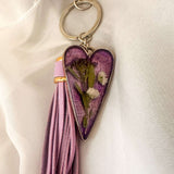 Antique silver Lavender Skies heart bag charm with a soft purple resin background and white dried flowers, attached to a matching purple leather tassel.