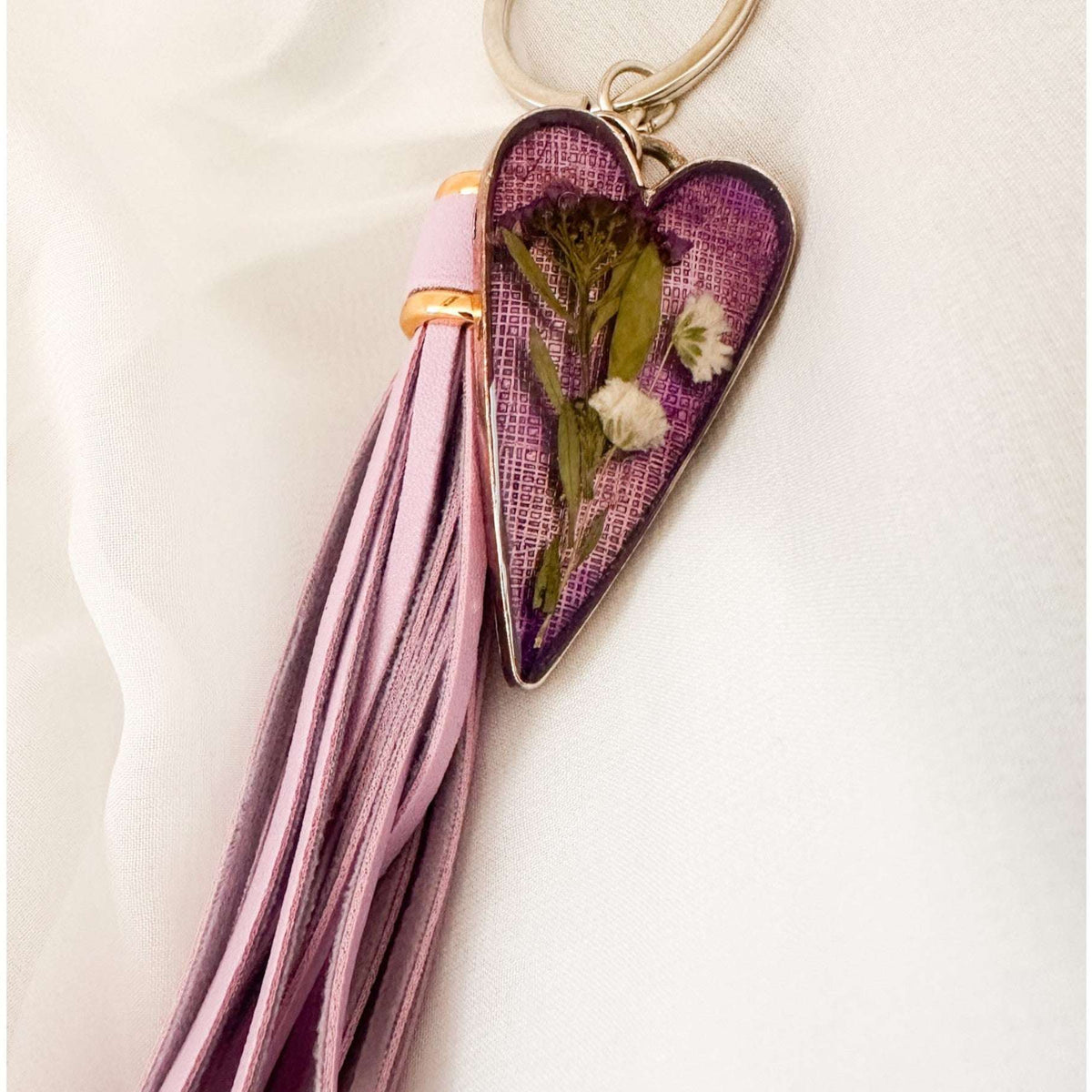 Antique silver Lavender Skies heart bag charm with a soft purple resin background and white dried flowers, attached to a matching purple leather tassel.ain with tassel and pressed flowers on a light background