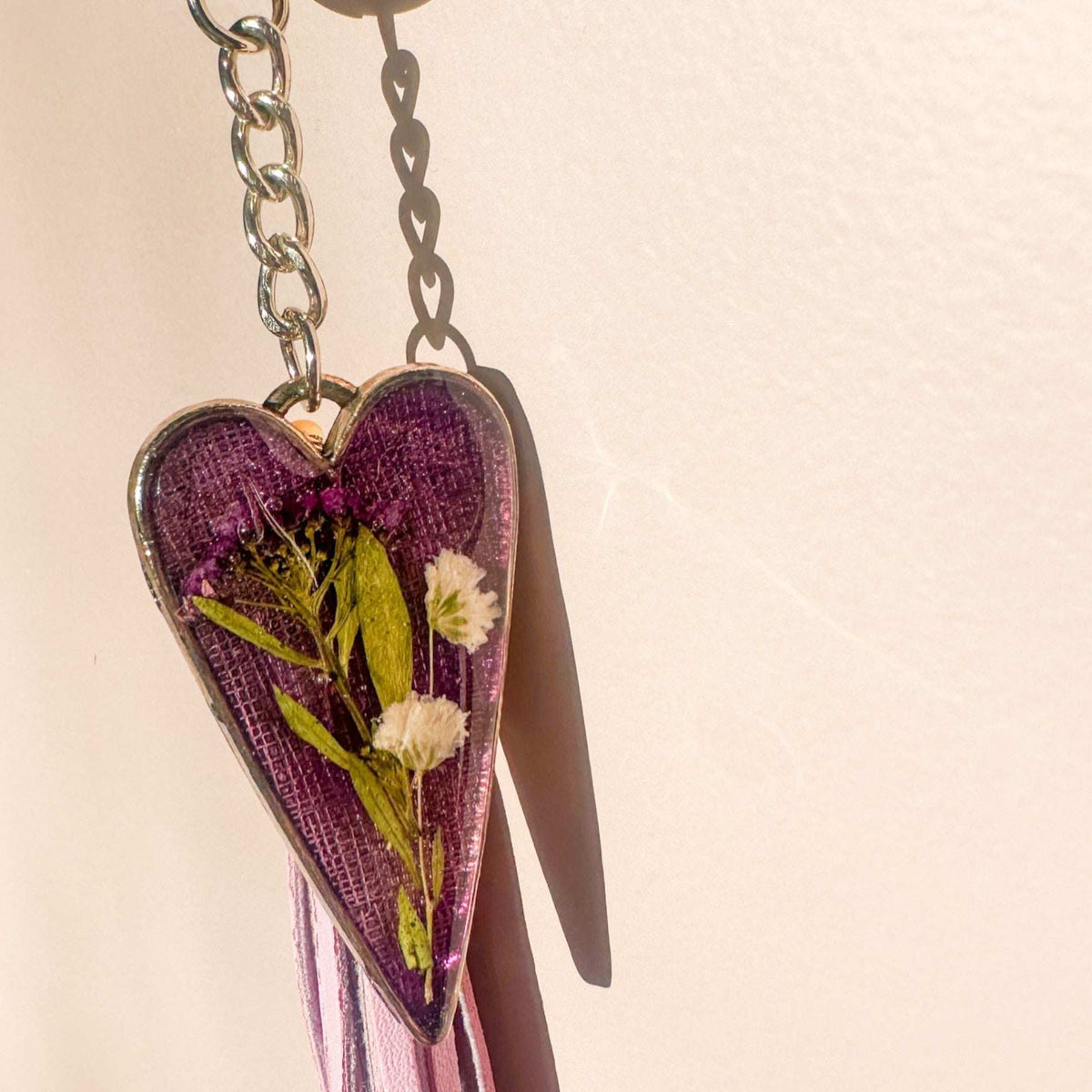 Antique silver heart Lavender Skies bag charm with a soft purple resin background and white dried flowers, attached to a matching purple leather tassel.with dried flowers on a beige background