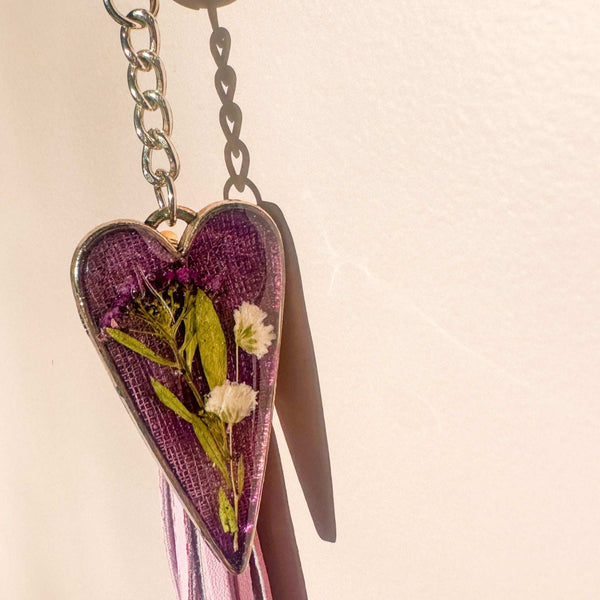 Antique silver heart Lavender Skies bag charm with a soft purple resin background and white dried flowers, attached to a matching purple leather tassel.with dried flowers on a beige background
