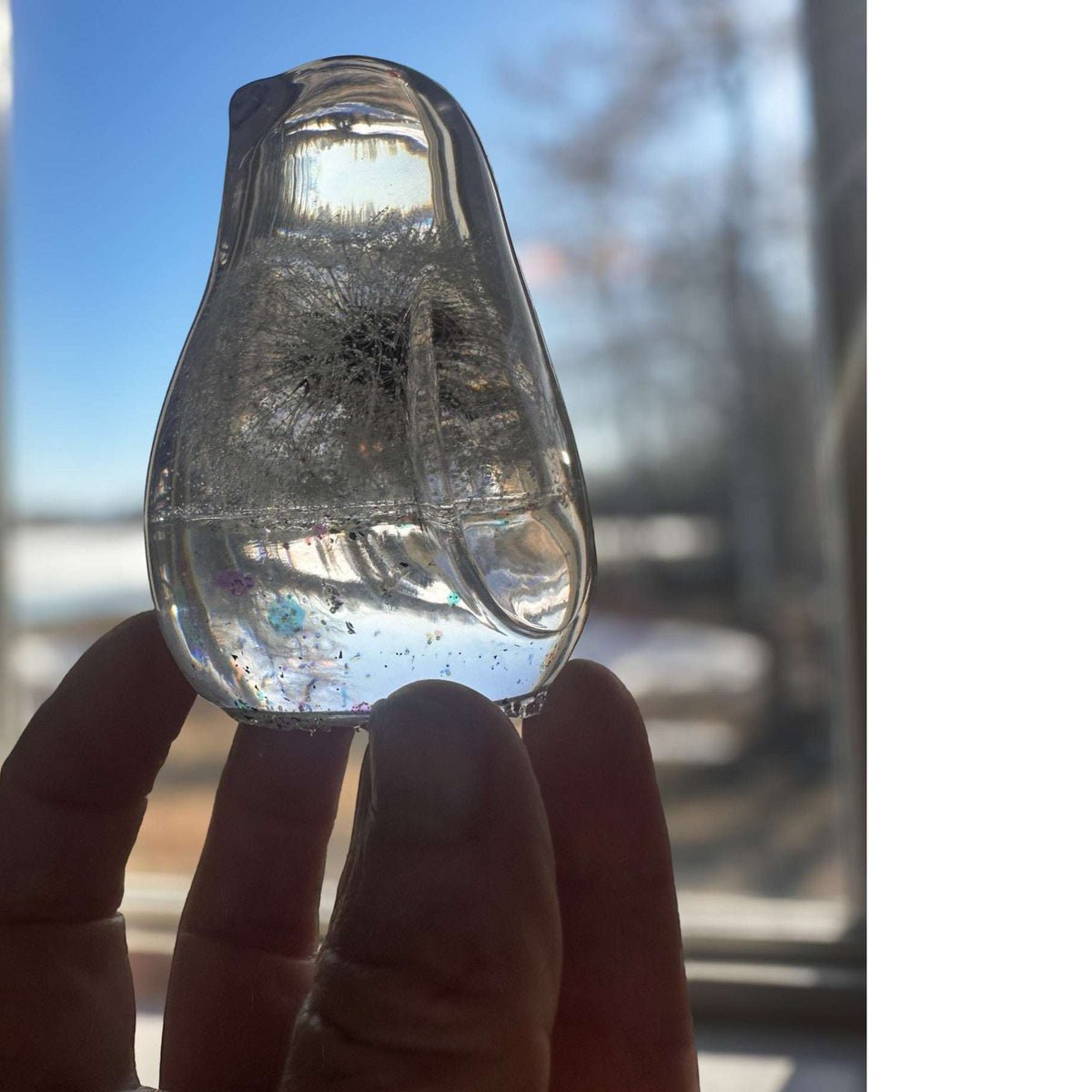 Close-up of preserved dandelion seed head suspended in clear hand-poured resin bird held against blue sky