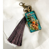 Handmade teal resin keychain with a silver fairy on real Boreal forest mushrooms and white wildflowers, brown tassel and antique copper hardware, white background.