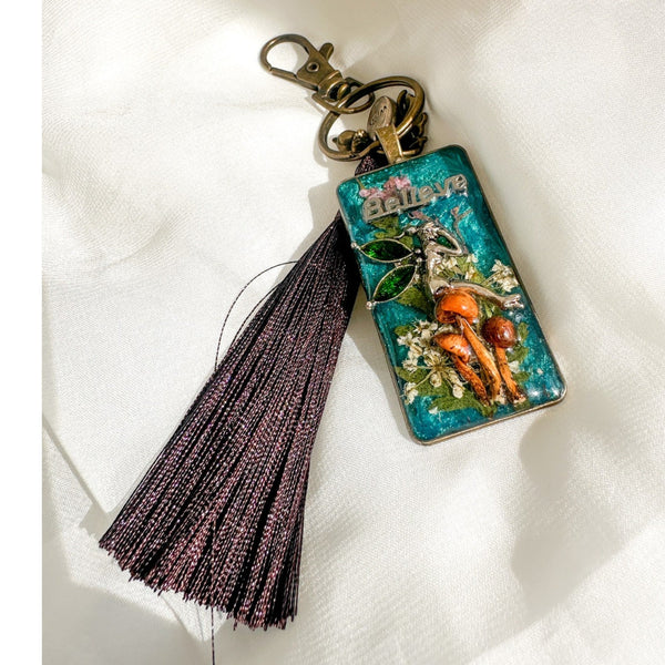 Handmade teal resin keychain with a silver fairy on real Boreal forest mushrooms and white wildflowers, brown tassel and antique copper hardware, white background.