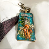 Handmade teal resin keychain with a silver fairy on real Boreal forest mushrooms and white wildflowers, brown tassel and antique copper hardware, white background.