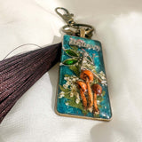 Handmade teal resin keychain with a silver fairy on real Boreal forest mushrooms and white wildflowers, brown tassel and antique copper hardware, white background.