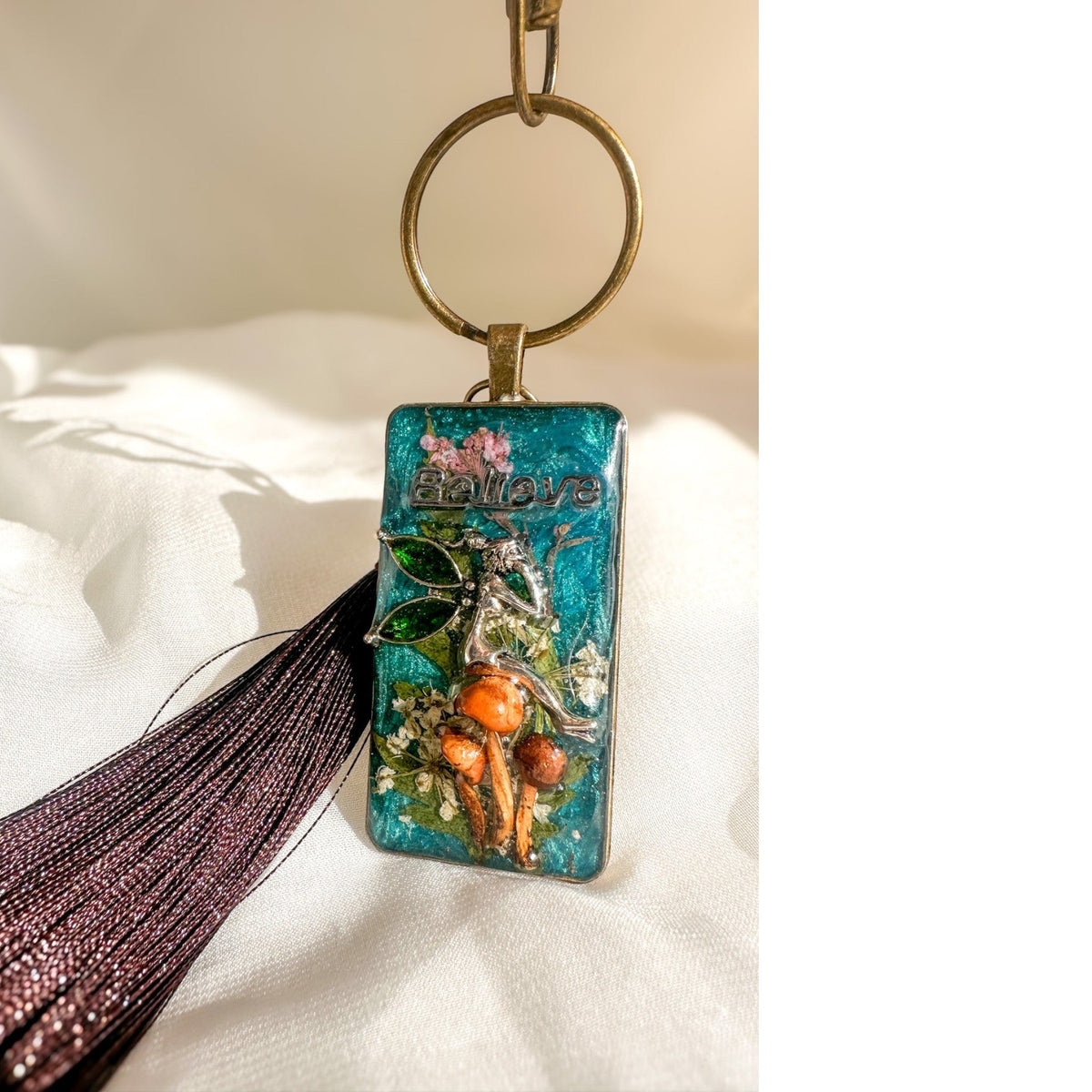 Handmade teal resin keychain with a silver fairy on real Boreal forest mushrooms and white wildflowers, brown tassel and antique copper hardware, white background.