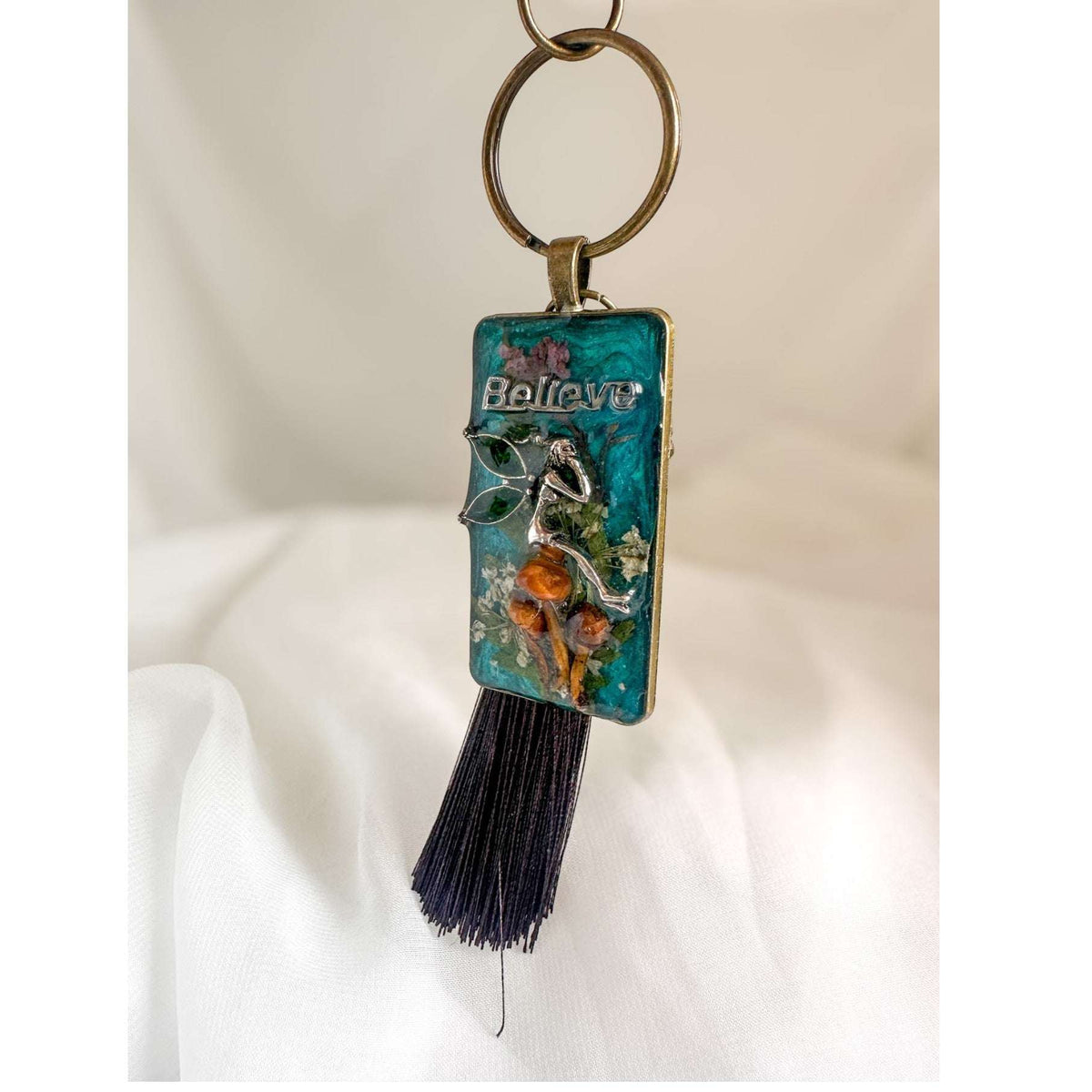 Handmade teal resin keychain with a silver fairy on real Boreal forest mushrooms and white wildflowers, brown tassel and antique copper hardware, white background.
