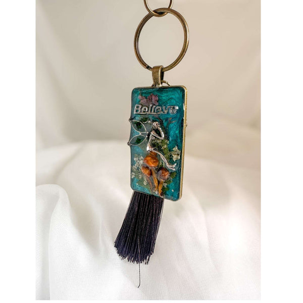 Handmade teal resin keychain with a silver fairy on real Boreal forest mushrooms and white wildflowers, brown tassel and antique copper hardware, white background.