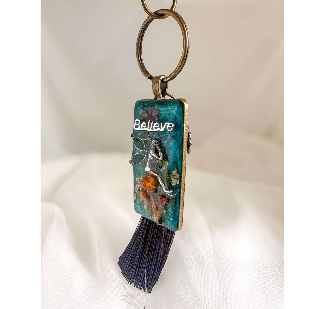 Handmade teal resin keychain with a silver fairy on real Boreal forest mushrooms and white wildflowers, brown tassel and antique copper hardware, white background.