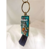 Handmade teal resin keychain with a silver fairy on real Boreal forest mushrooms and white wildflowers, brown tassel and antique copper hardware, white background.