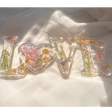 Daisy resin word artwith pressed white daisy, blush blooms, gold leaf flakes, and heart-shaped O design