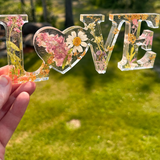Daisy resin word art with real pressed white daisy and blush blooms, gold leaf flakes, heart-shaped O, and purple wildflower accents.