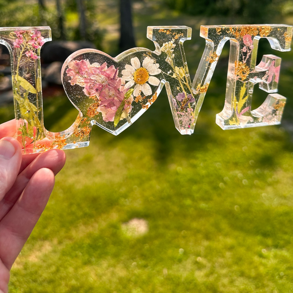 Daisy resin word art with real pressed white daisy and blush blooms, gold leaf flakes, heart-shaped O, and purple wildflower accents.