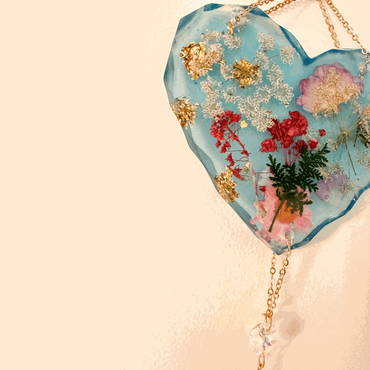 Glow in the dark heart suncatcher in blue resin with real pressed wildflowers, gold leaf accents, and a gold chain with crystal drops.
