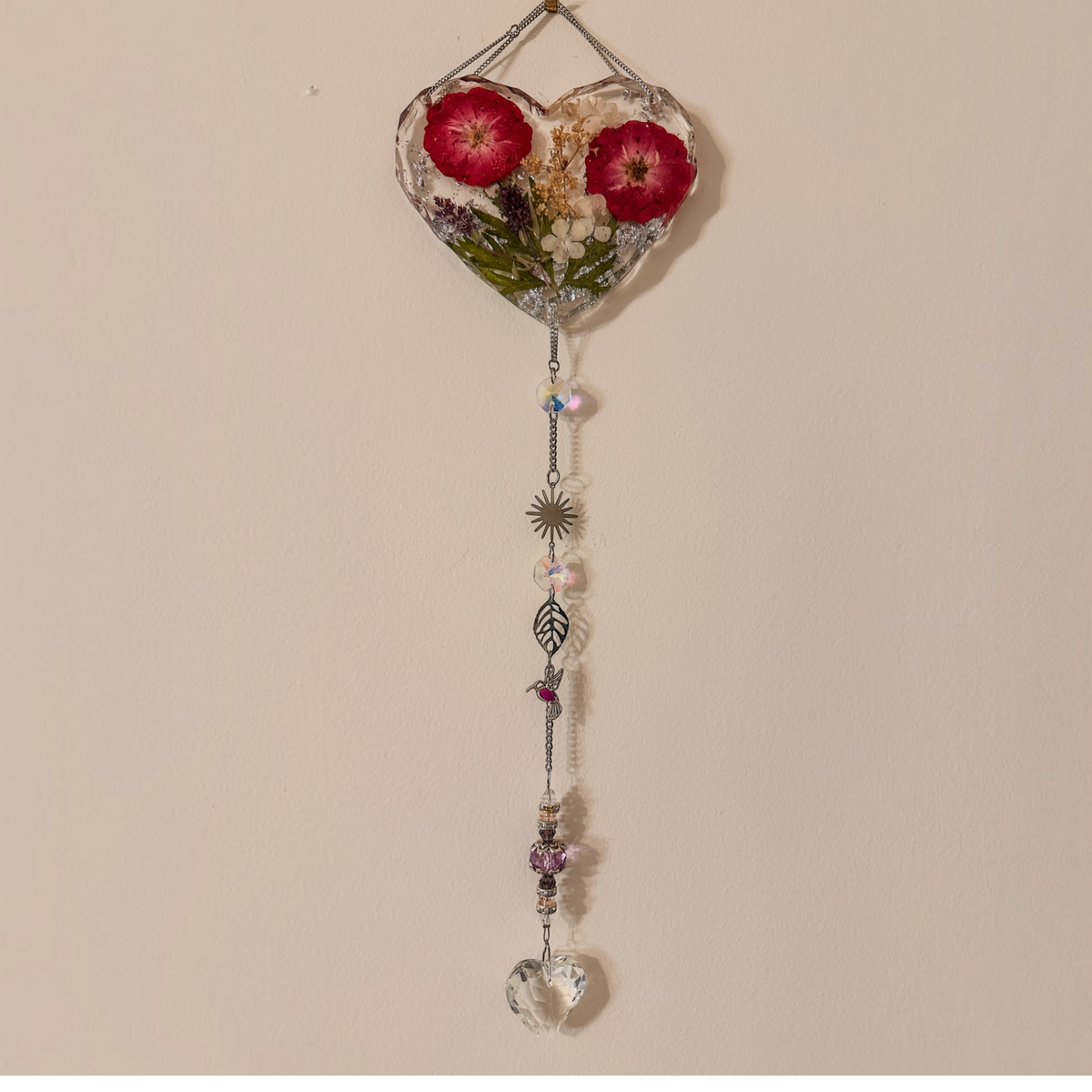 Pressed rose suncatcher featuring two deep crimson roses in clear resin with silver leaf, hanging silver chain with crystal prisms, sunburst, leaf, and hummingbird charms.