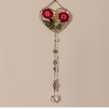 Pressed rose suncatcher featuring two deep crimson roses in clear resin with silver leaf, hanging silver chain with crystal prisms, sunburst, leaf, and hummingbird charms.