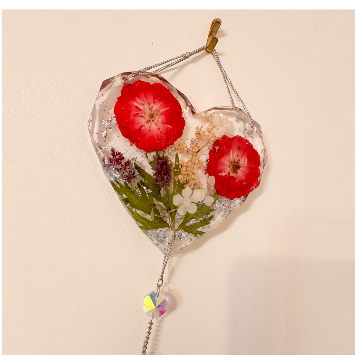 Heart-shaped clear resin pressed rose suncatcher with two red roses, green leaves, white and purple flowers, silver flakes, and hanging crystal prism.