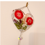 Heart-shaped clear resin pressed rose suncatcher with two red roses, green leaves, white and purple flowers, silver flakes, and hanging crystal prism.