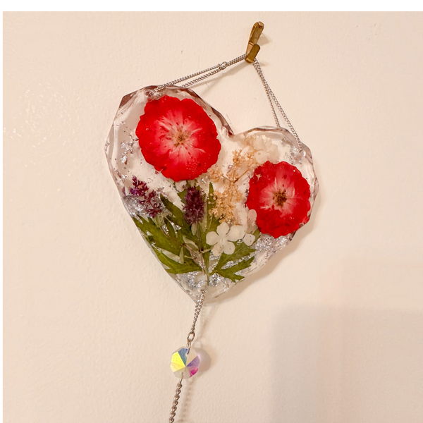 Heart-shaped clear resin pressed rose suncatcher with two red roses, green leaves, white and purple flowers, silver flakes, and hanging crystal prism.