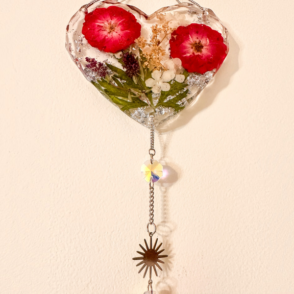 Heart-shaped clear resin pressed rose suncatcher with two deep red flowers, green leaves, silver flakes, and hanging crystal prisms and sunburst charm.