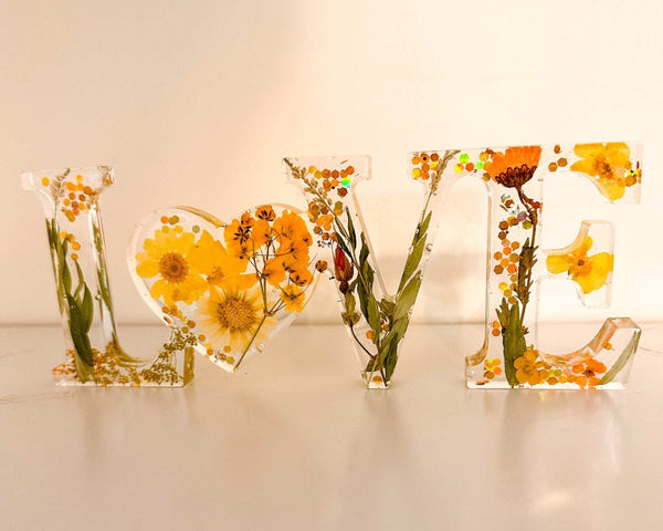 Pressed flower resin love sign in clear resin, featuring yellow wildflowers, greenery, and a floral heart-shaped O