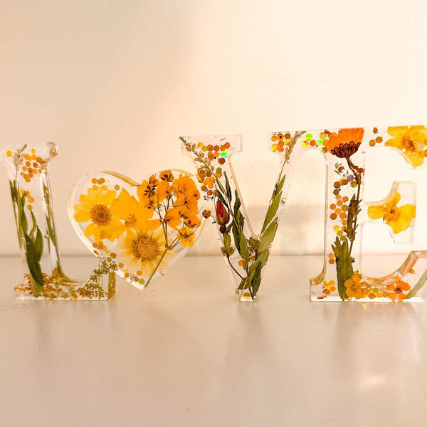 Pressed flower resin love sign in clear resin, featuring yellow wildflowers, greenery, and a floral heart-shaped O
