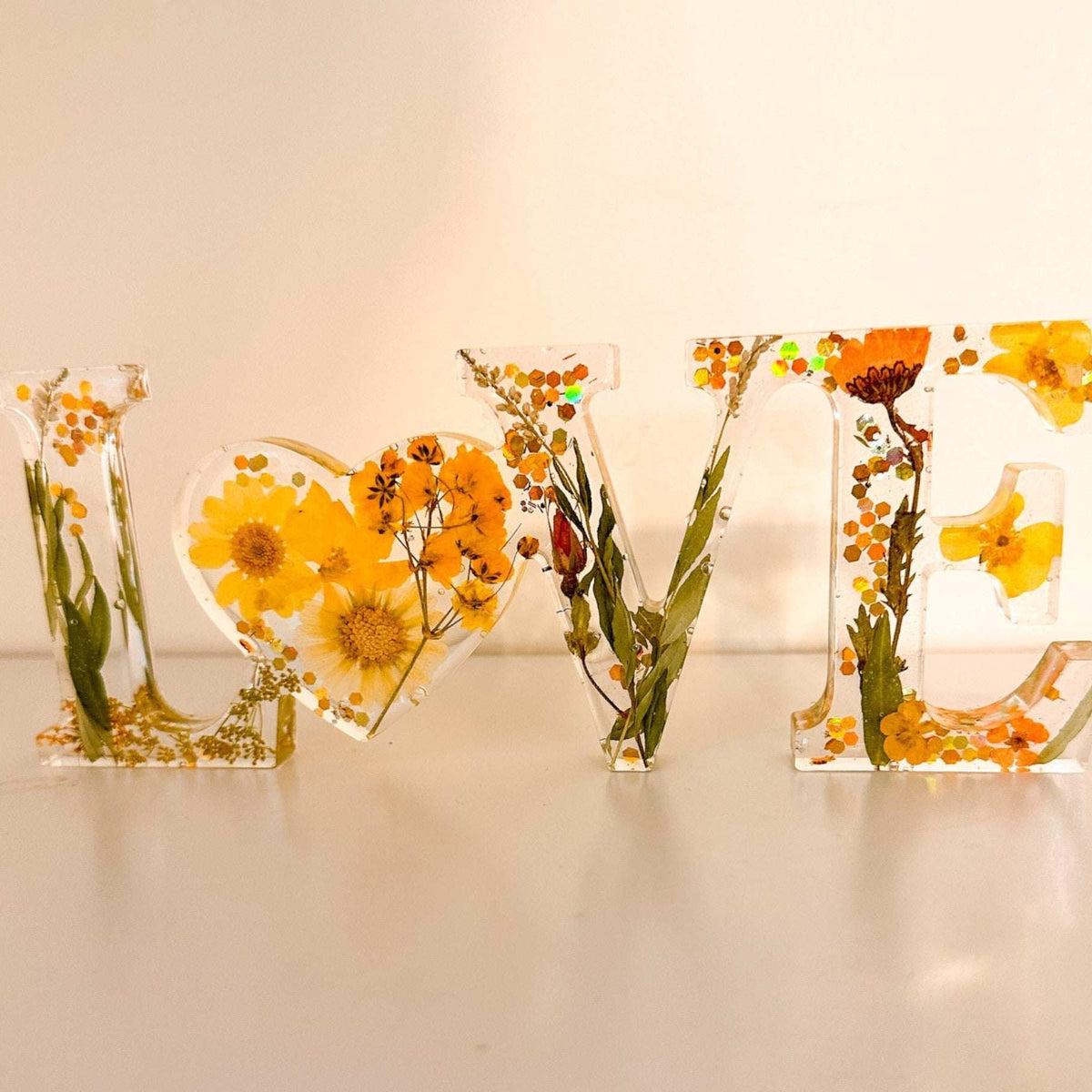 Pressed flower resin love sign in clear resin with yellow wildflowers, greenery, and a heart-shaped O centerpiece