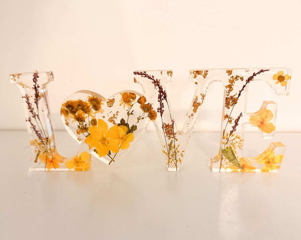 Pressed flower resin love sign with clear freestanding letters filled with yellow wildflowers and greenery, heart-shaped O