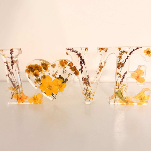 Pressed flower resin love sign with clear freestanding letters filled with yellow wildflowers and greenery, heart-shaped O