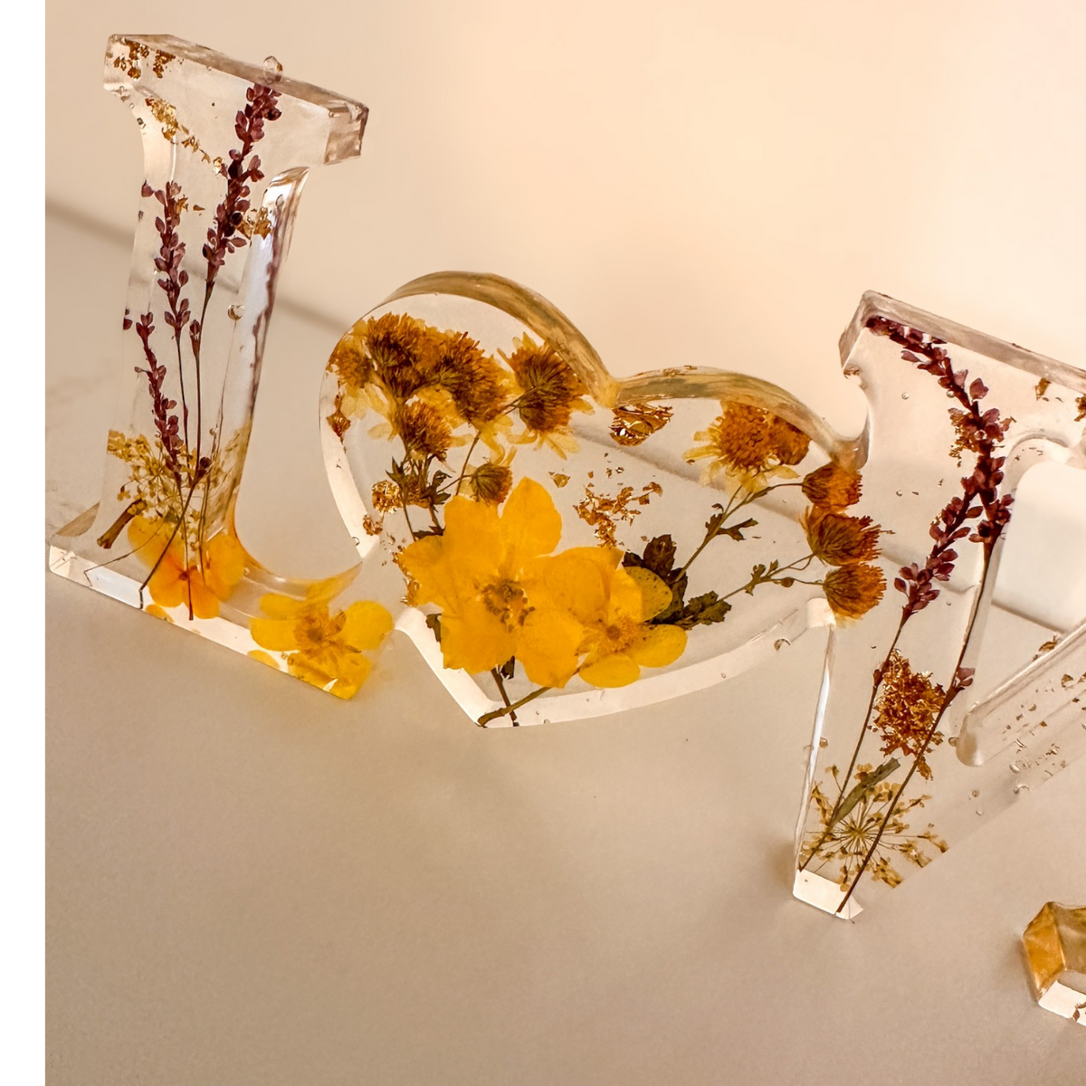 Pressed flower resin love sign with clear letters and a heart-shaped O filled with yellow dried wildflowers and greenery