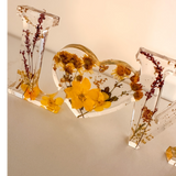 Pressed flower resin love sign with clear letters and a heart-shaped O filled with yellow dried wildflowers and greenery