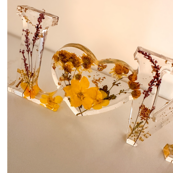 Pressed flower resin love sign with clear letters and a heart-shaped O filled with yellow dried wildflowers and greenery