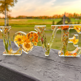 Pressed flower resin love sign with clear letters, yellow blooms, orange flowers, greenery, and a heart-shaped O centerpiece