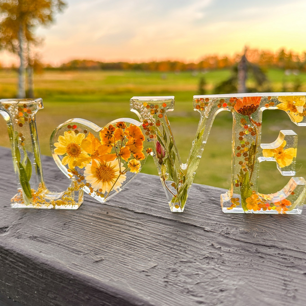 Pressed flower resin love sign with clear letters, yellow blooms, orange flowers, greenery, and a heart-shaped O centerpiece