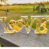 Pressed flower resin love sign in clear resin with yellow wildflowers, greenery, and a heart-shaped O centerpiece