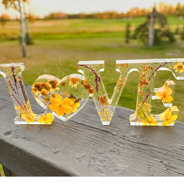 Pressed flower resin love sign in clear resin with yellow wildflowers, greenery, and a heart-shaped O centerpiece