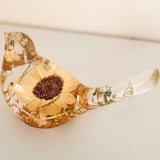Resin bird decor with a pressed Sunflower, golden-brown tones, delicate green botanicals, and a clear glossy finish.