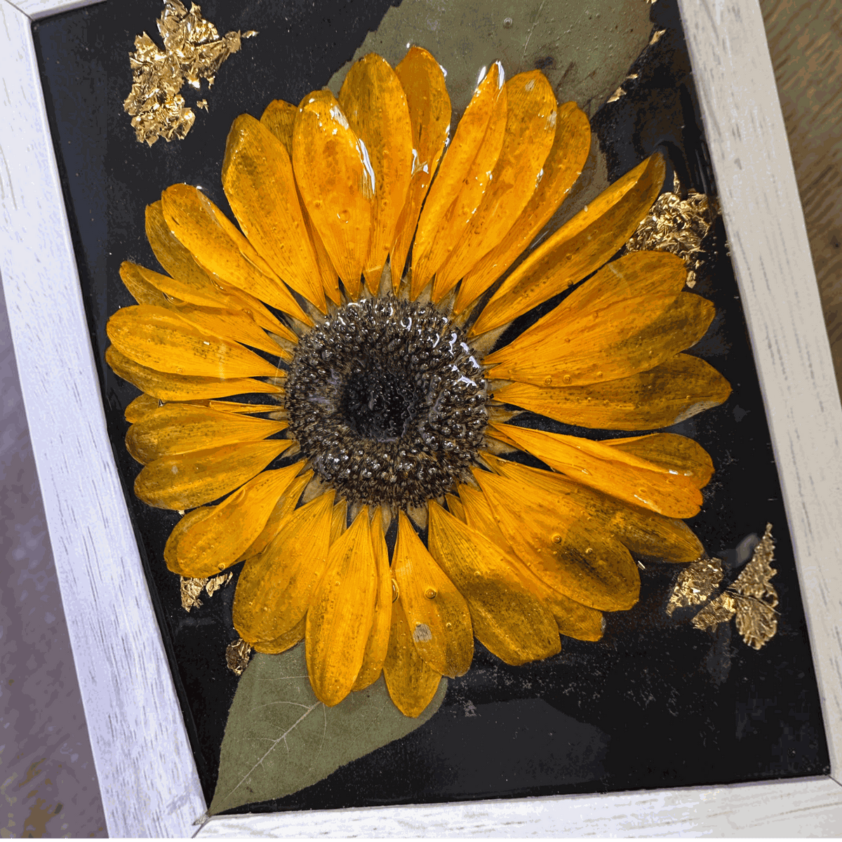 Pressed sunflower frame art with a dimensional orange bloom, green leaves, gold flecks on black, and a pale wood frame.