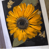 Pressed sunflower frame art with a dimensional orange bloom, green leaves, gold flecks on black, and a pale wood frame.
