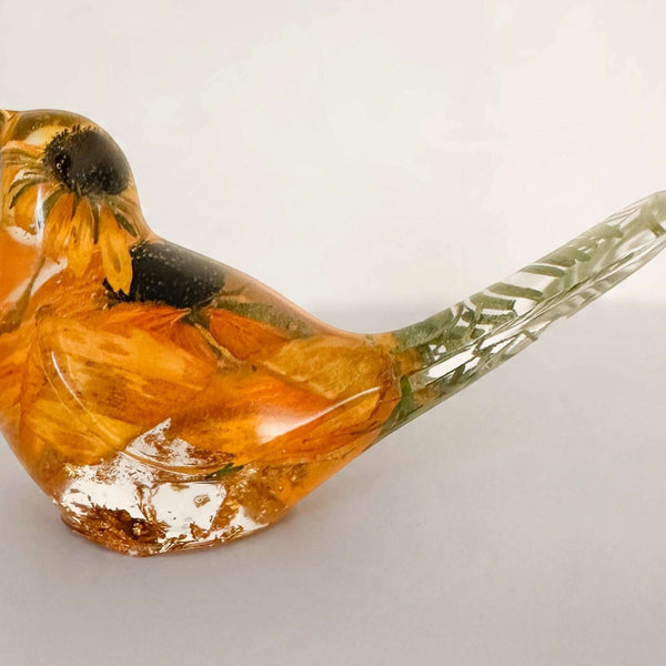 Resin bird in clear amber-toned resin with a preserved golden flower, dark centre, and flecks of gold leaf; Golden Meadow decor.
