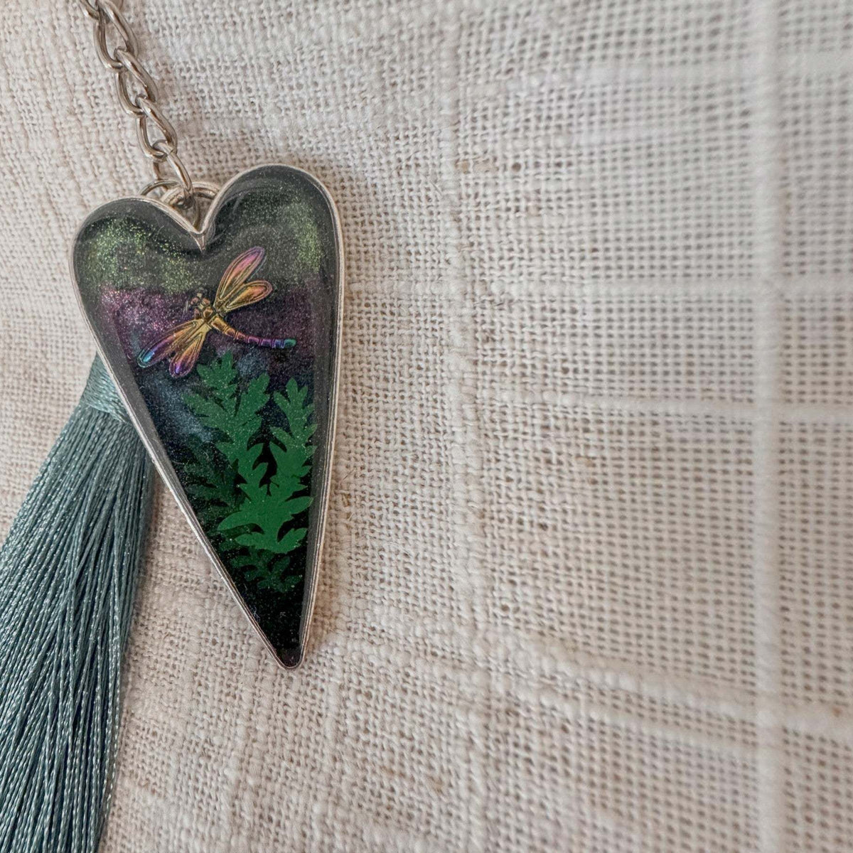 Aurora dragonfly keychain in a silver heart frame, with green ferns, shimmering purple-green resin, and a teal tassel.