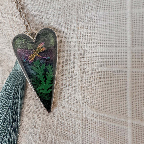Aurora dragonfly keychain in a silver heart frame, with green ferns, shimmering purple-green resin, and a teal tassel.