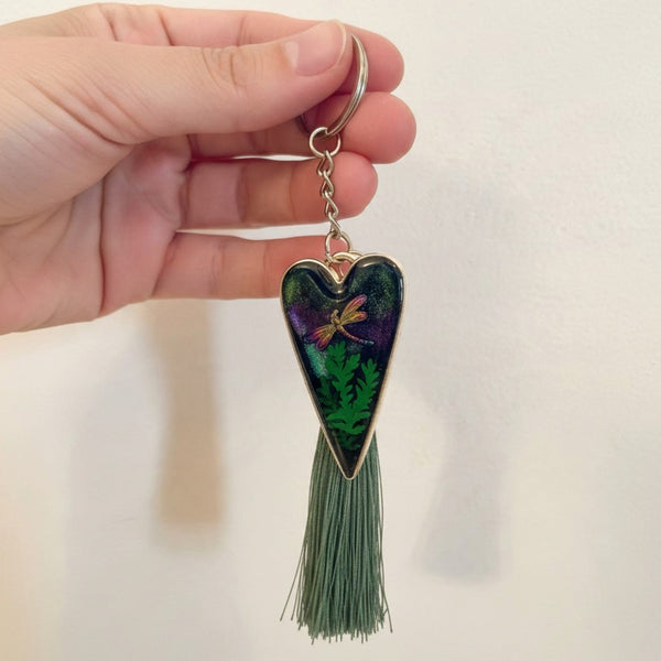 Aurora dragonfly keychain with a silver heart frame, dark green and purple resin, fern pattern, dragonfly and green tassel.
