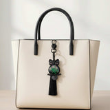 Bag Charm featuring a dark owl frame, green and purple shimmering resin body, and long black tassel on a cream handbag