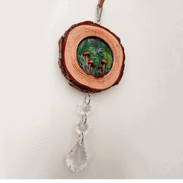 boreal forest suncatcher on a natural bark-edged wood slice, with a green mushroom scene, dragonfly and hanging clear crystals