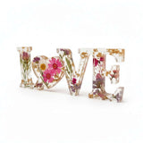 Wildflower resin word art spelling LOVE with pink daisies, white daisy, purple wildflowers, rose buds, greenery, and gold leaf flakes