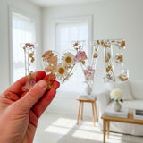 Whispering Meadow LOVE resin word art with white daisies, pink and purple wildflowers, bunny tail accent, and gold leaf flakes.