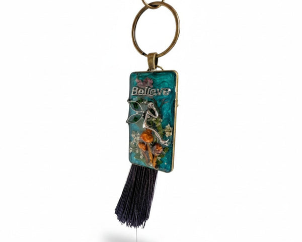 Handmade teal resin keychain with a silver fairy on real Boreal forest mushrooms and white wildflowers, brown tassel and antique copper hardware, white background.
