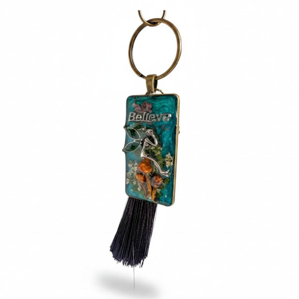 Handmade teal resin keychain with a silver fairy on real Boreal forest mushrooms and white wildflowers, brown tassel and antique copper hardware, white background.