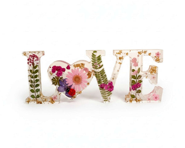 Pink daisy resin word art with pressed wildflowers, tiny ladybug, fern fronds, and gold leaf flakes in clear resin letters spelling LOVE